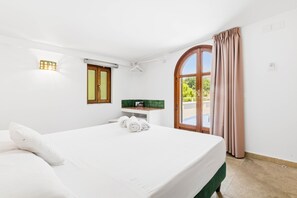 A hotel room with a bed, a small table, a green cushioned bench, a window with curtains, and a view of greenery outside.