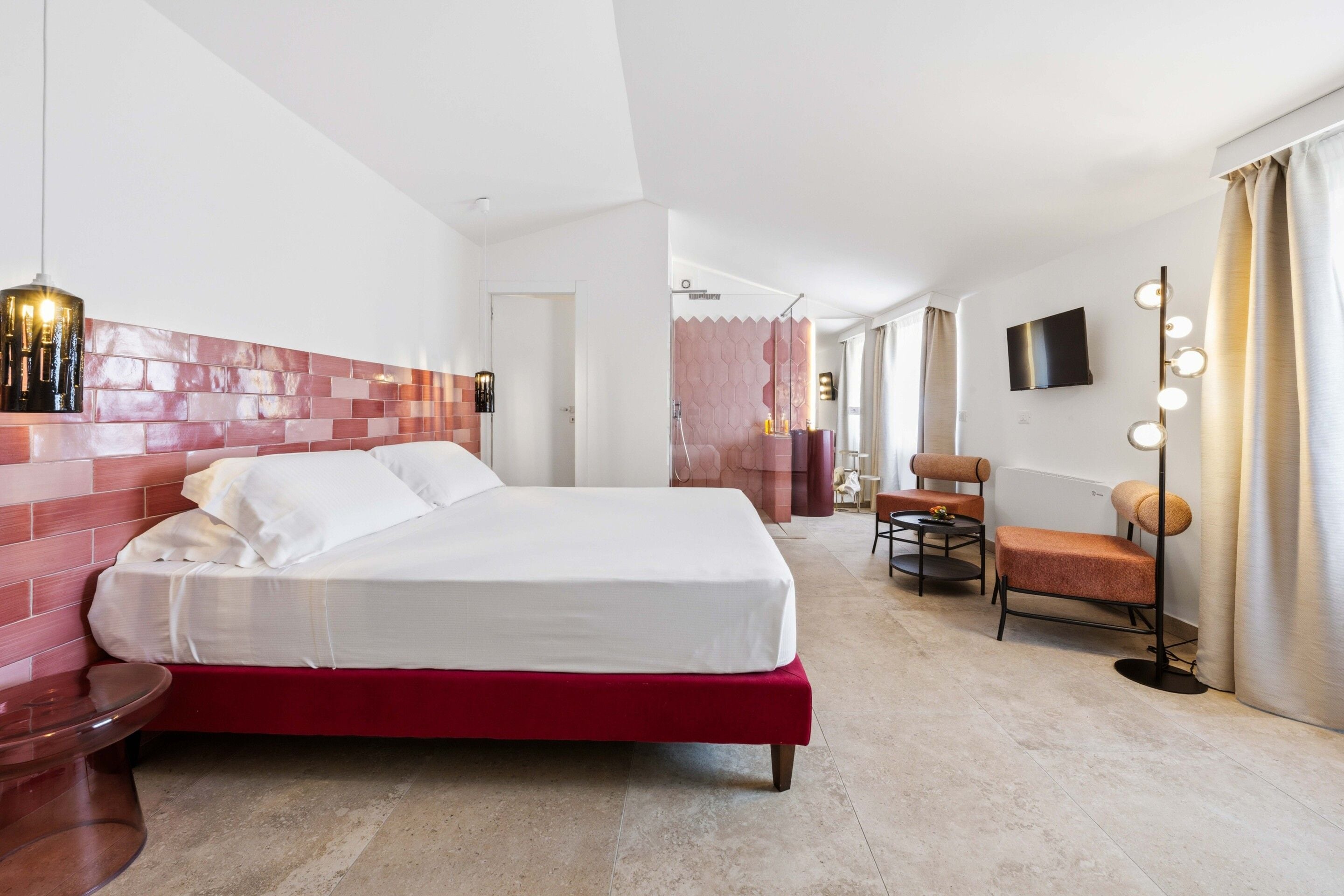 Habitación de hotel con cama, zona de estar con una silla y una mesa, televisor montado en la pared y espejo con luces.