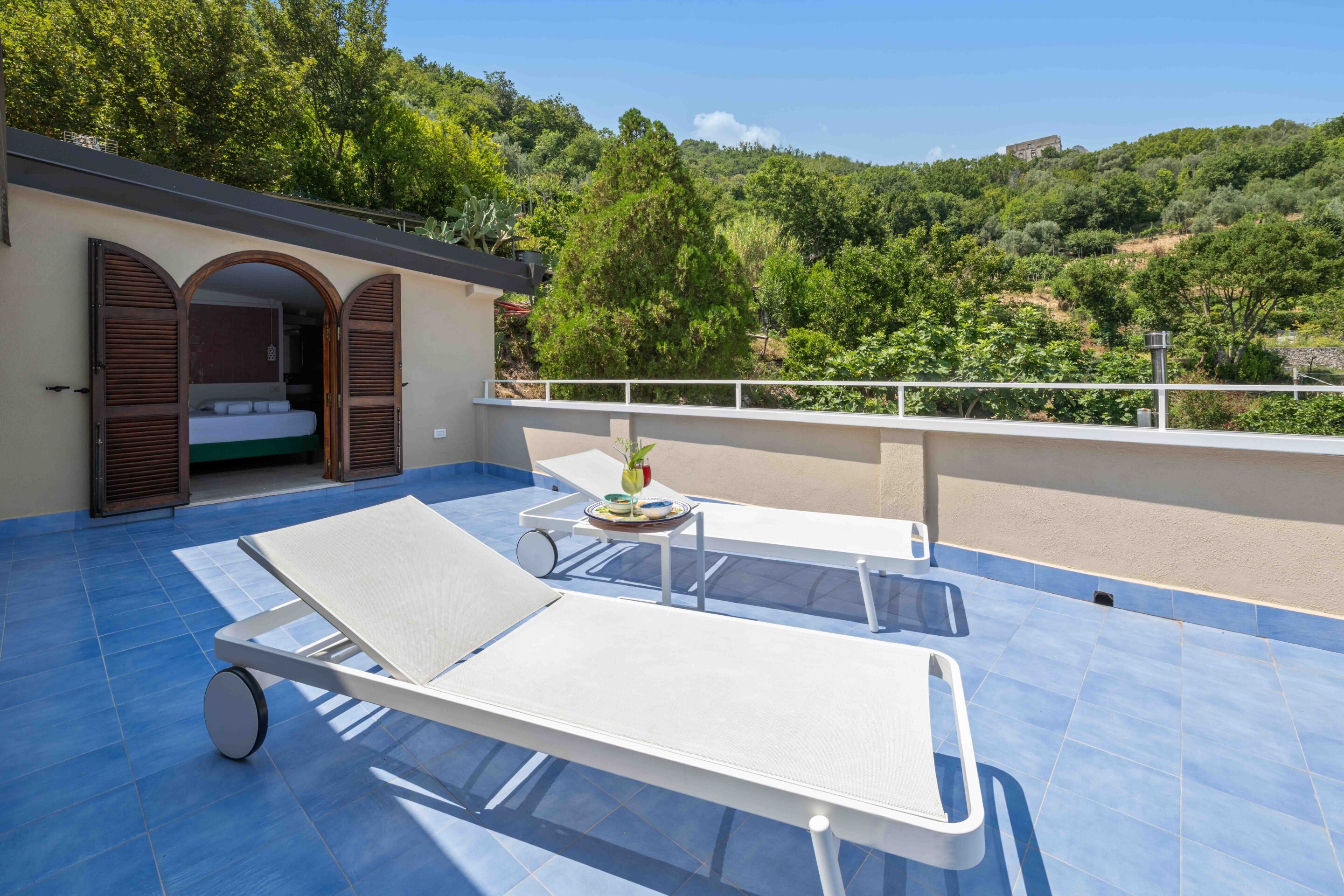 A rooftop terrace with two lounge chairs, a small table, and a view of a forested hillside.