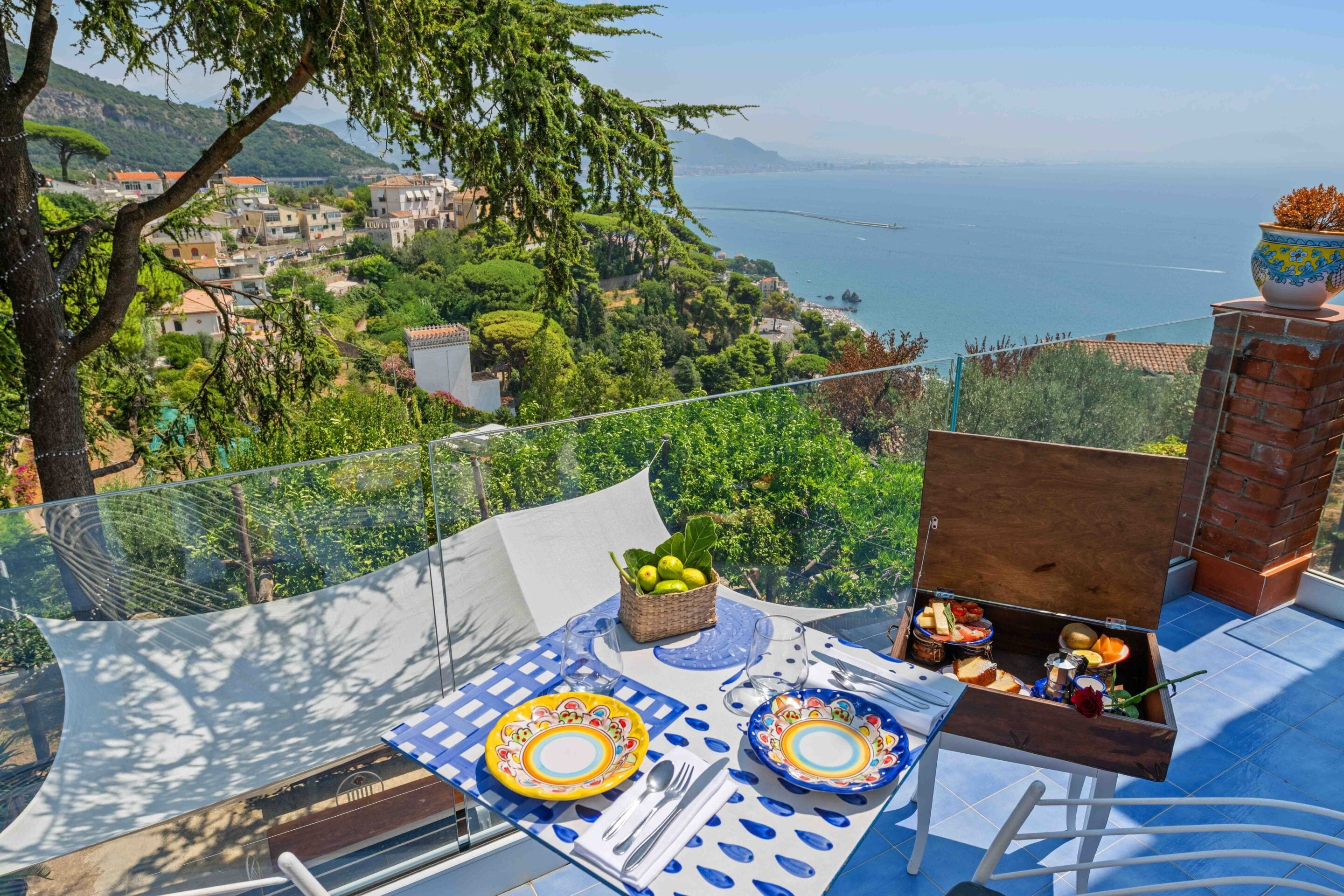 A terrace with a table set for dining, overlooking a coastal landscape with houses and a sea view.