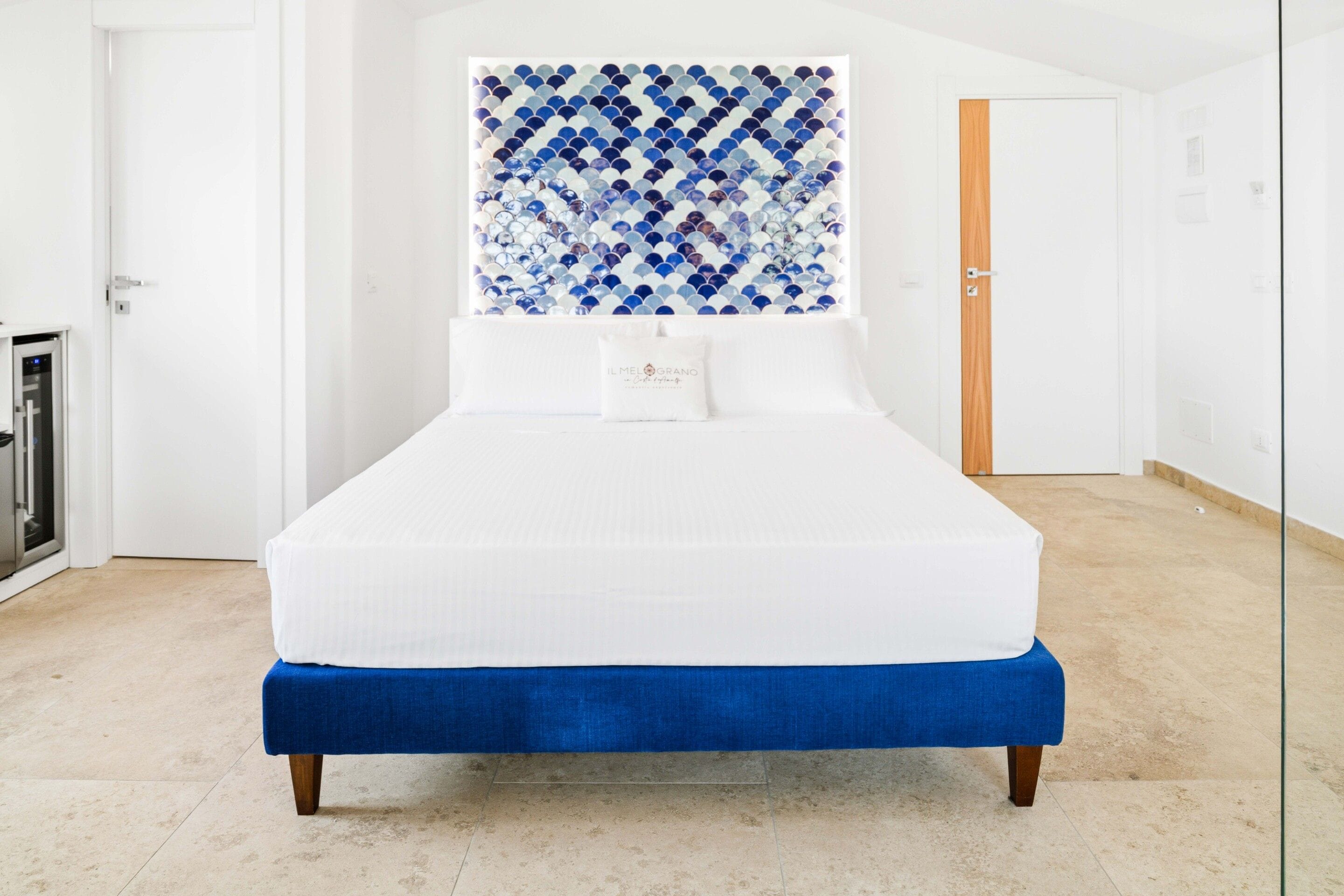 Una cama con colchón blanco y ropa de cama azul, una pared decorativa con azulejos azules y blancos, y una puerta de madera al fondo.