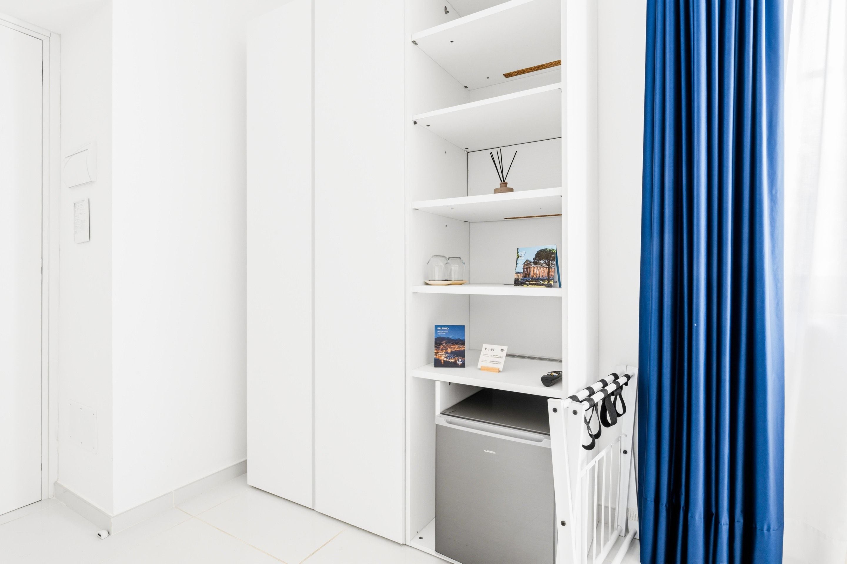 Una habitación blanca con un mini refrigerador empotrado, un estante con un libro y el símbolo de Wi-Fi, y una cortina azul.