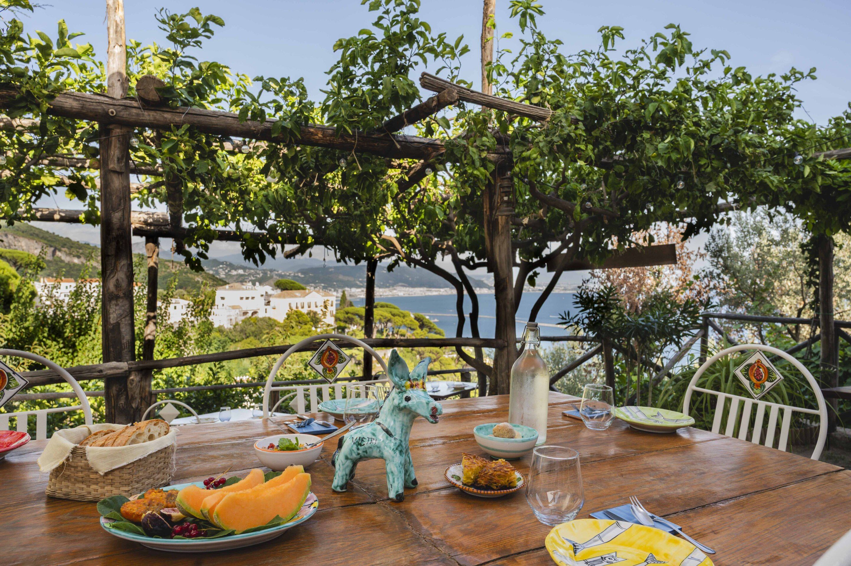 An outdoor dining table with food and a decorative horse statue, overlooking a scenic coastal view.