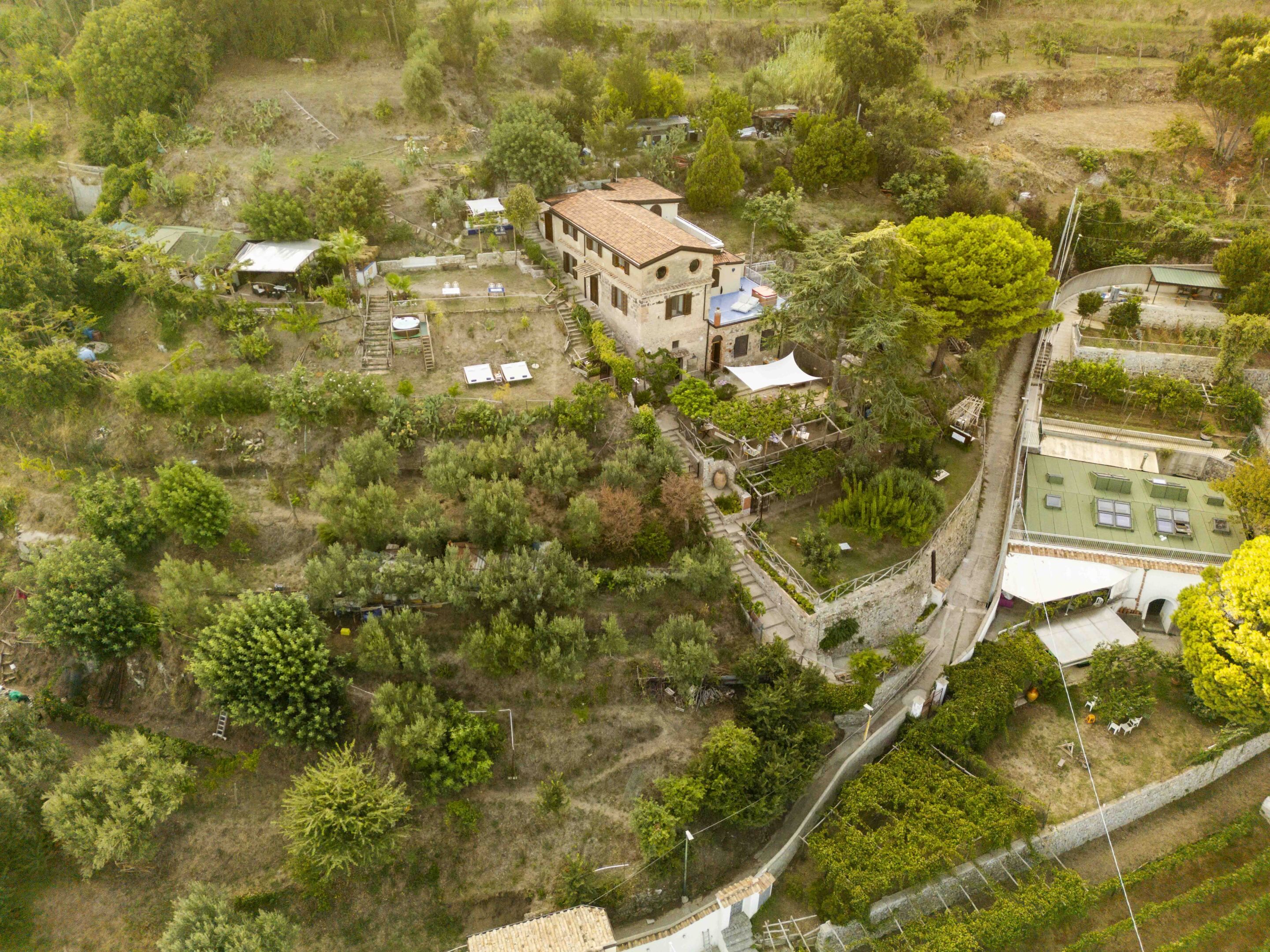 Vista aérea de una propiedad con un edificio central rodeado de vegetación, una zona pavimentada y una estructura pequeña a la derecha.