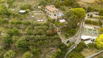 Vista aérea de una propiedad con un edificio central rodeado de vegetación, una zona pavimentada y una estructura pequeña a la derecha.