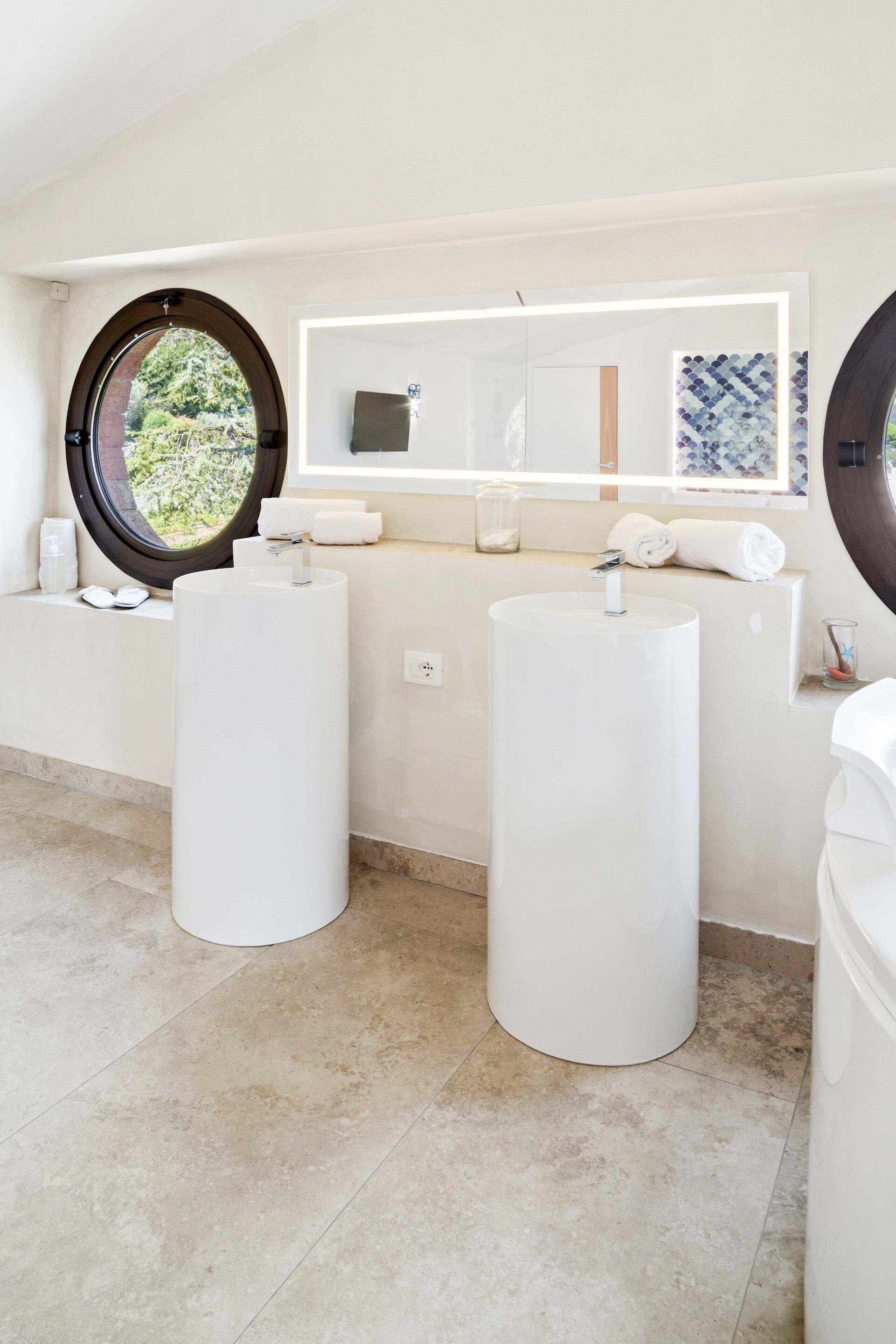 A modern bathroom with two large cylindrical sinks, a large mirror, and a window with a view of greenery.