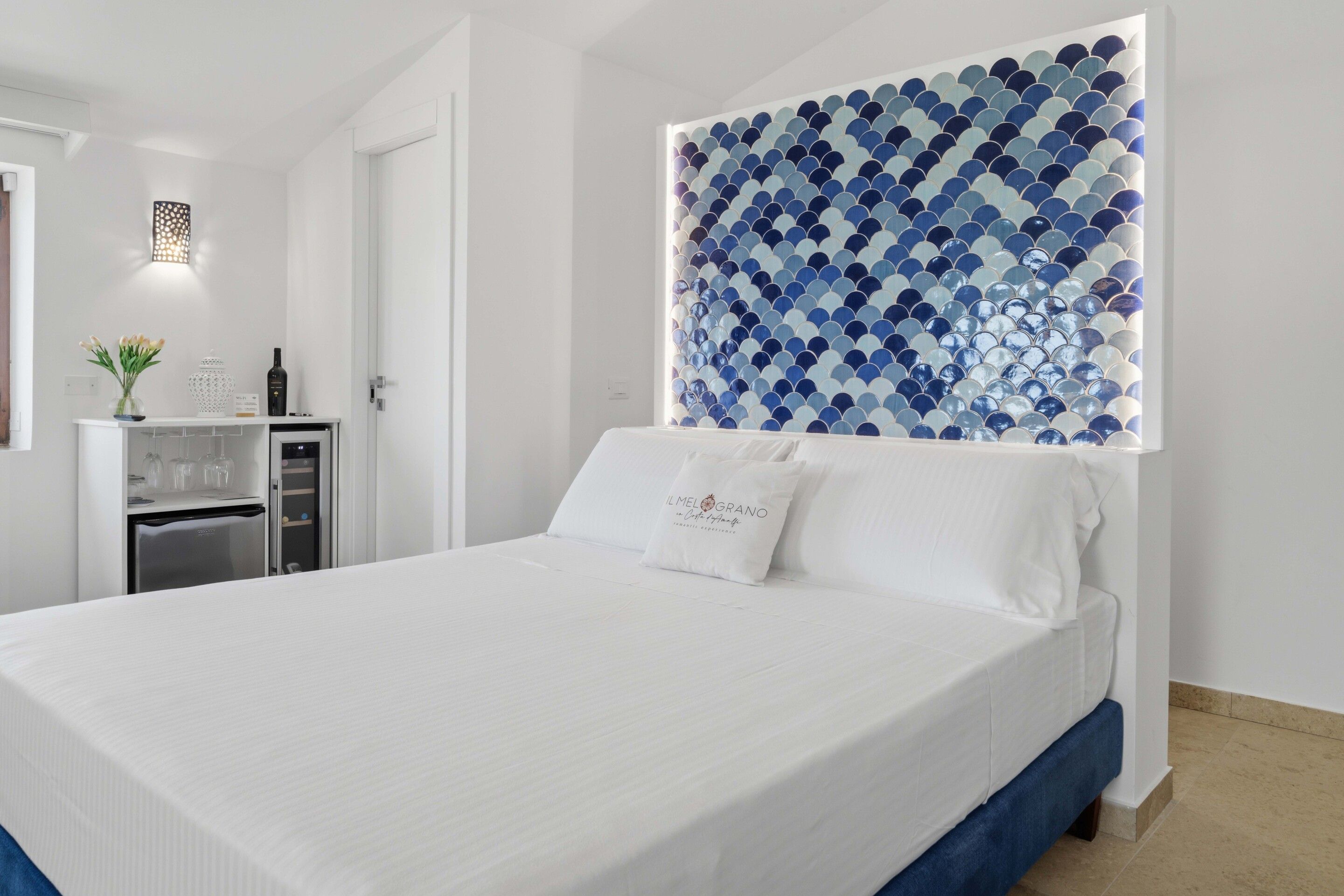 A hotel room with a bed, a minibar, a wall-mounted light fixture, and a decorative wall panel.