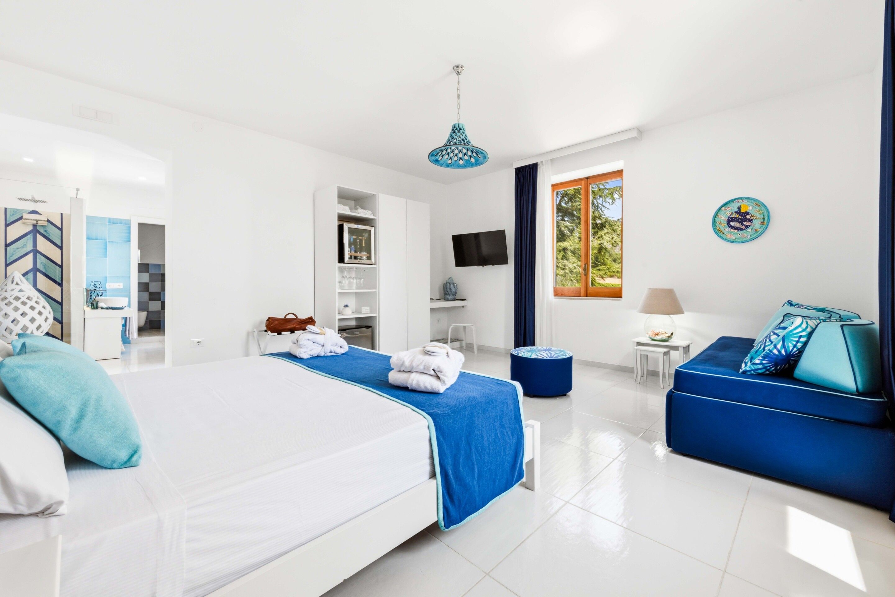 A modern hotel room with a large bed, a sofa, a TV, and a window with a view of trees.
