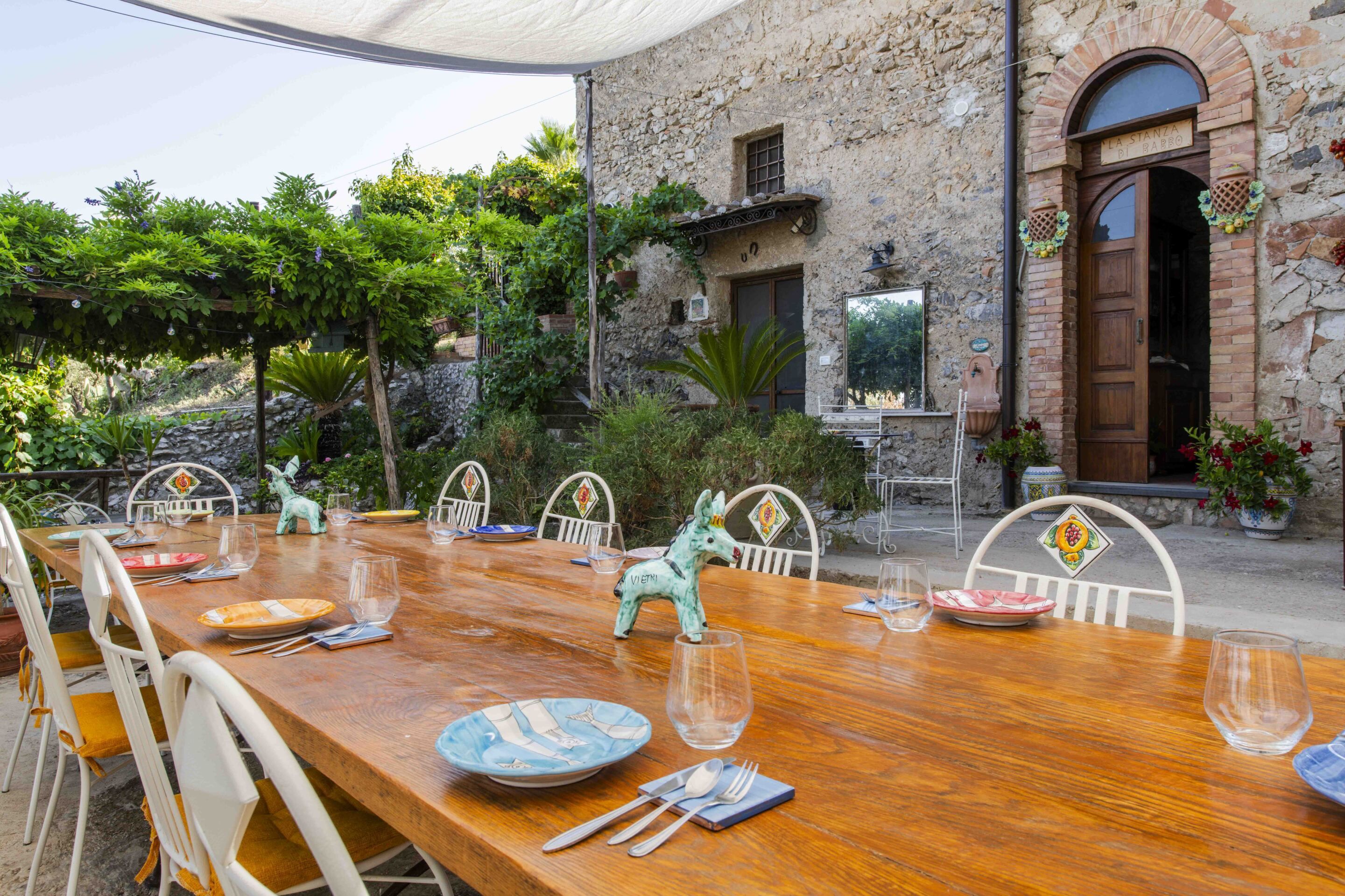 An outdoor dining area with a wooden table set for four, surrounded by stone walls and greenery.