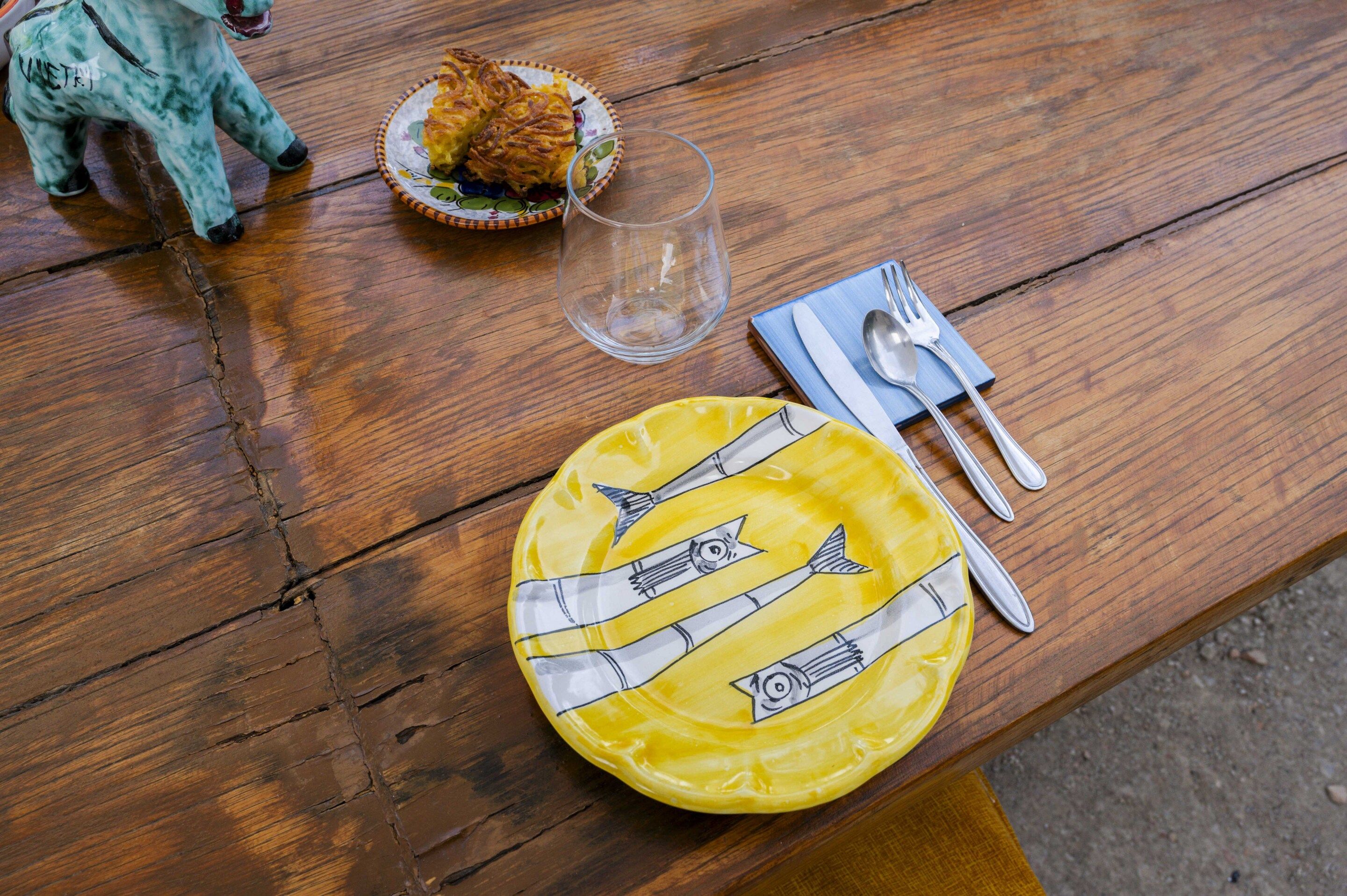 Una mesa de madera con un plato amarillo con ilustraciones de pescado, un vaso transparente, una pequeña figurita y una servilleta con cubiertos.