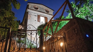 A gated entrance with wrought iron gates, a stone wall, and a building with a rounded tower and windows in the background.