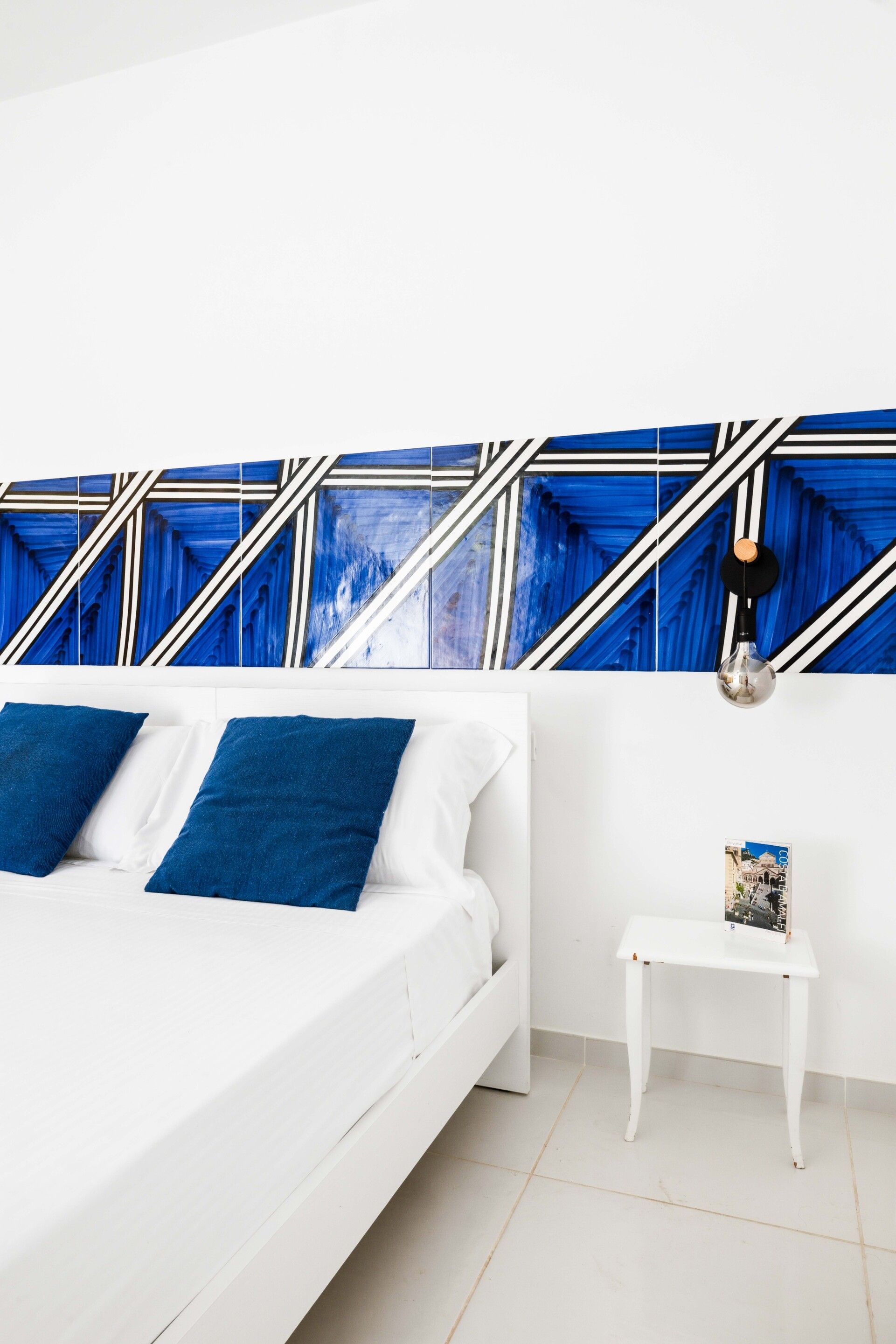 Un dormitorio con cama blanca, almohadas azules, una mesita de noche blanca y una pared con patrones geométricos azules y blancos.