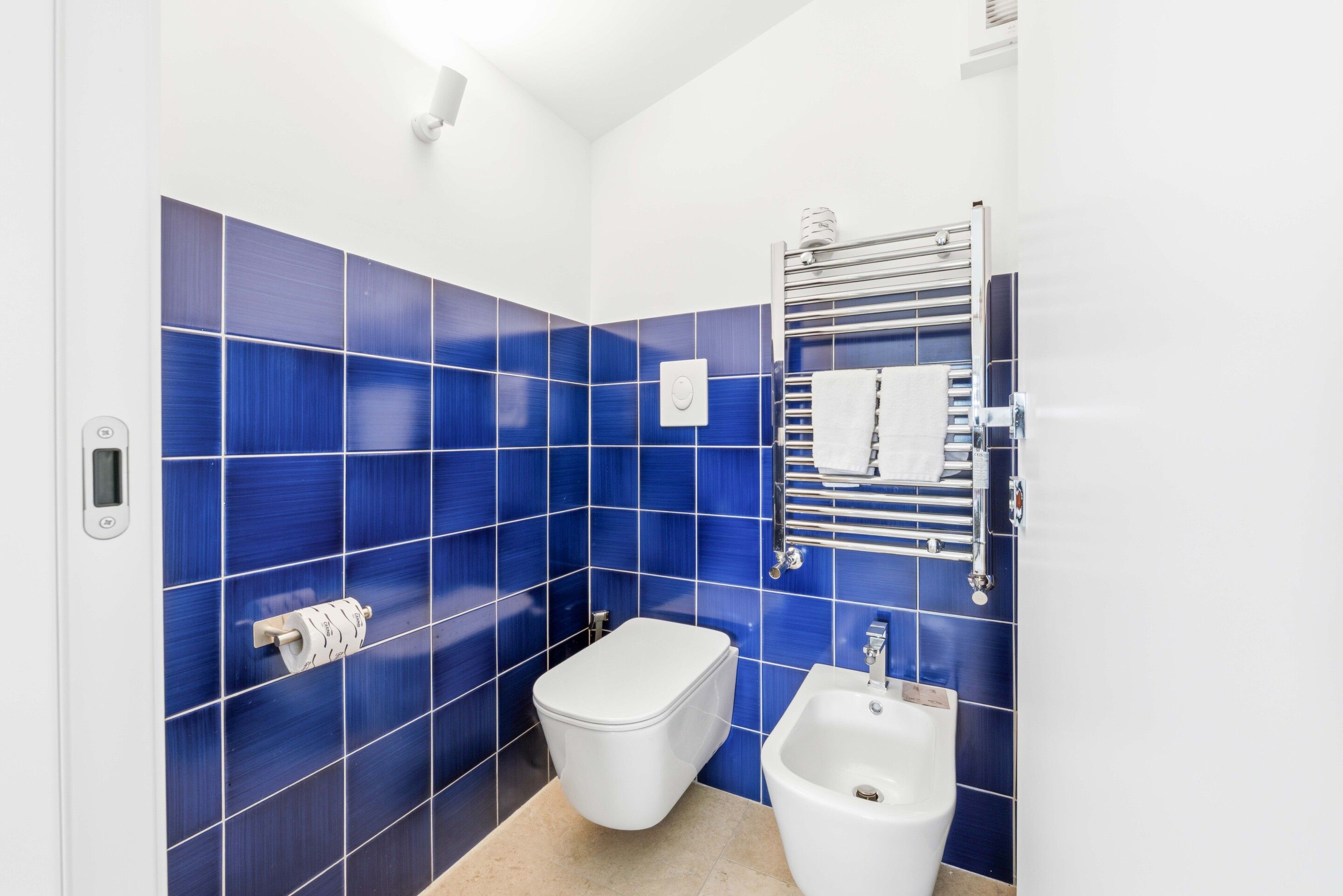A small, modern bathroom with blue tiles, a toilet, a bidet, and a towel rack.