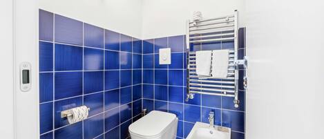 A small, modern bathroom with blue tiles, a toilet, a bidet, and a towel rack.