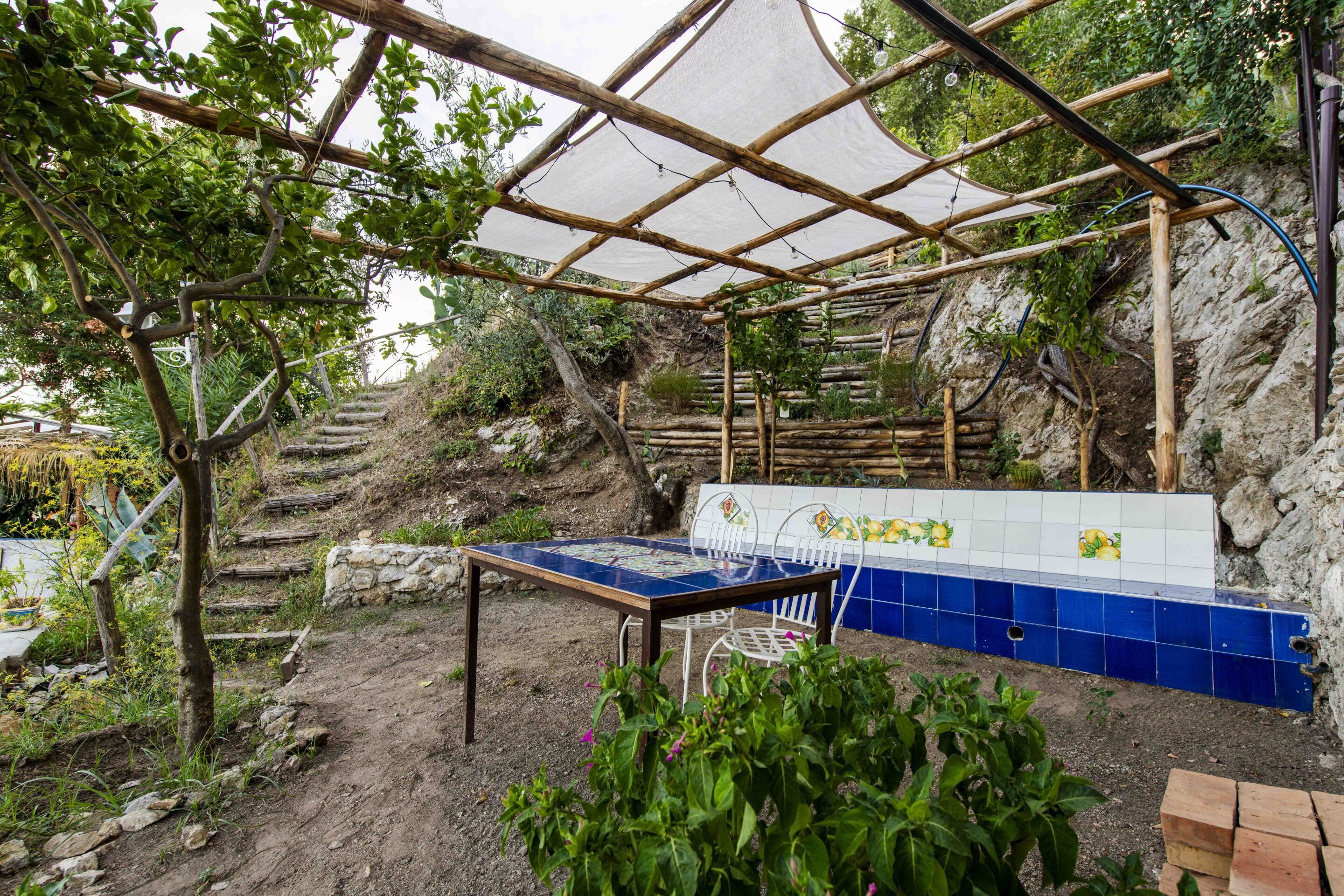 Un jardín al aire libre con una mesa y sillas, rodeado de plantas y un muro de piedra.