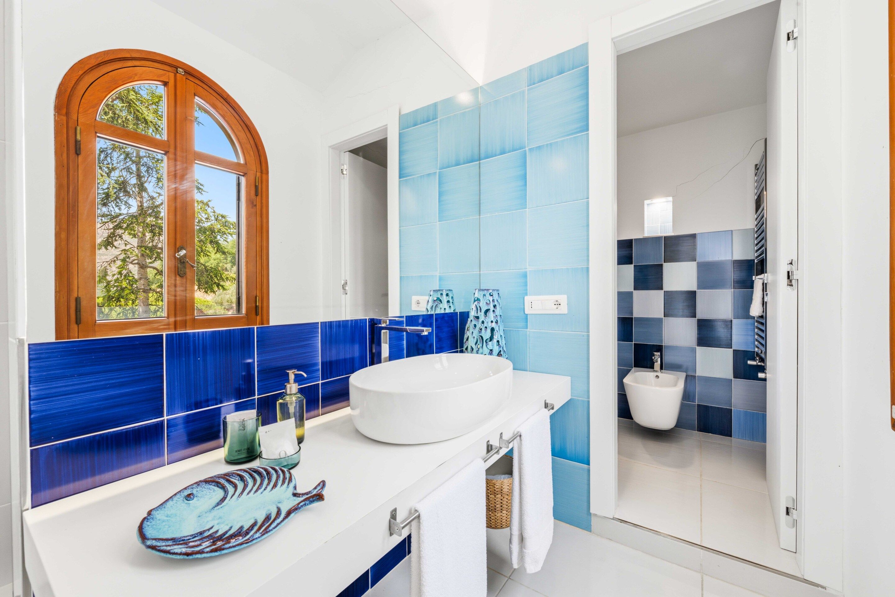 A bathroom with a white sink, blue tiles, a wooden-framed window, and a shower area.