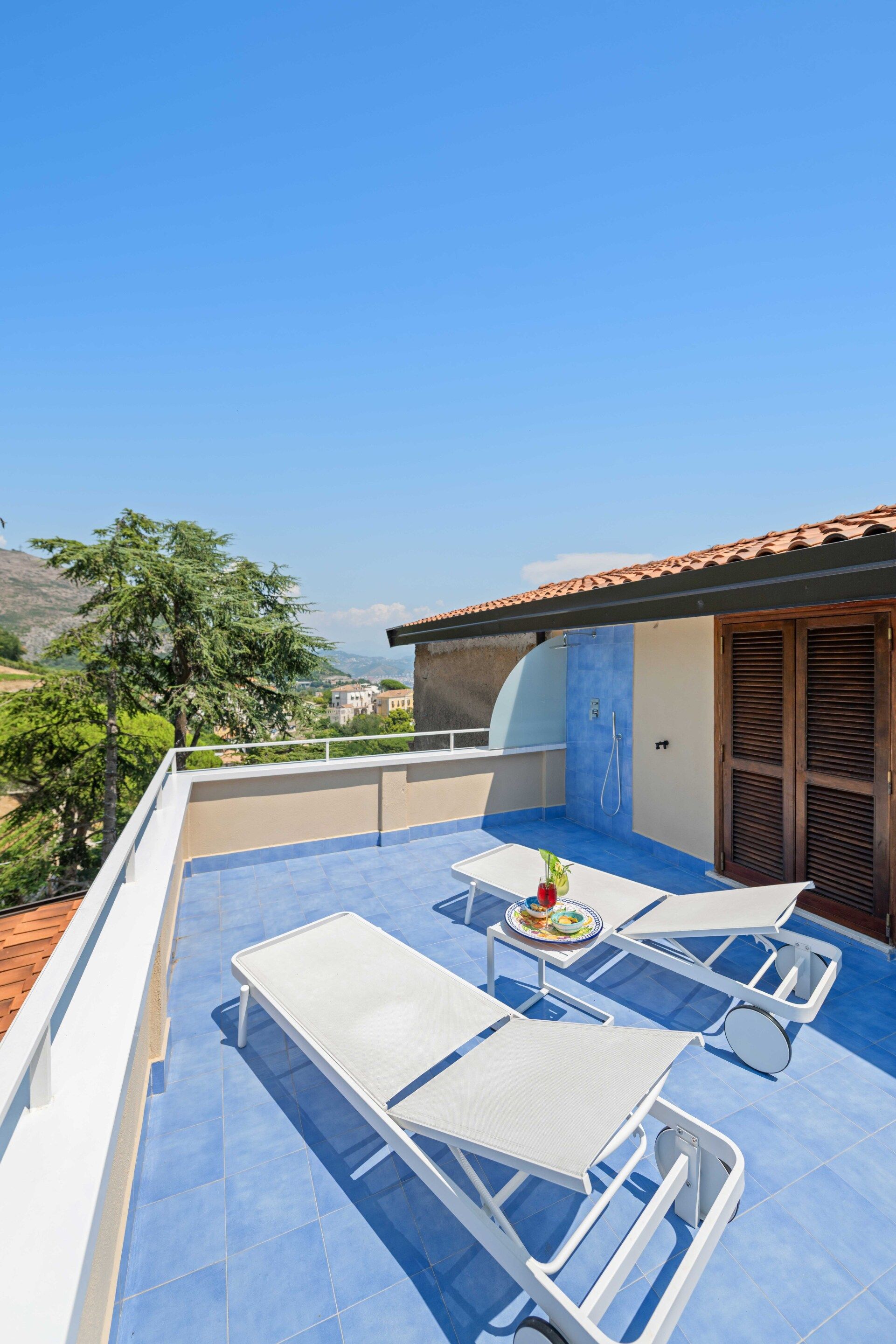 Una terraza en la azotea con dos sillones blancos, una mesita con un plato de comida y un suelo de baldosas azules.