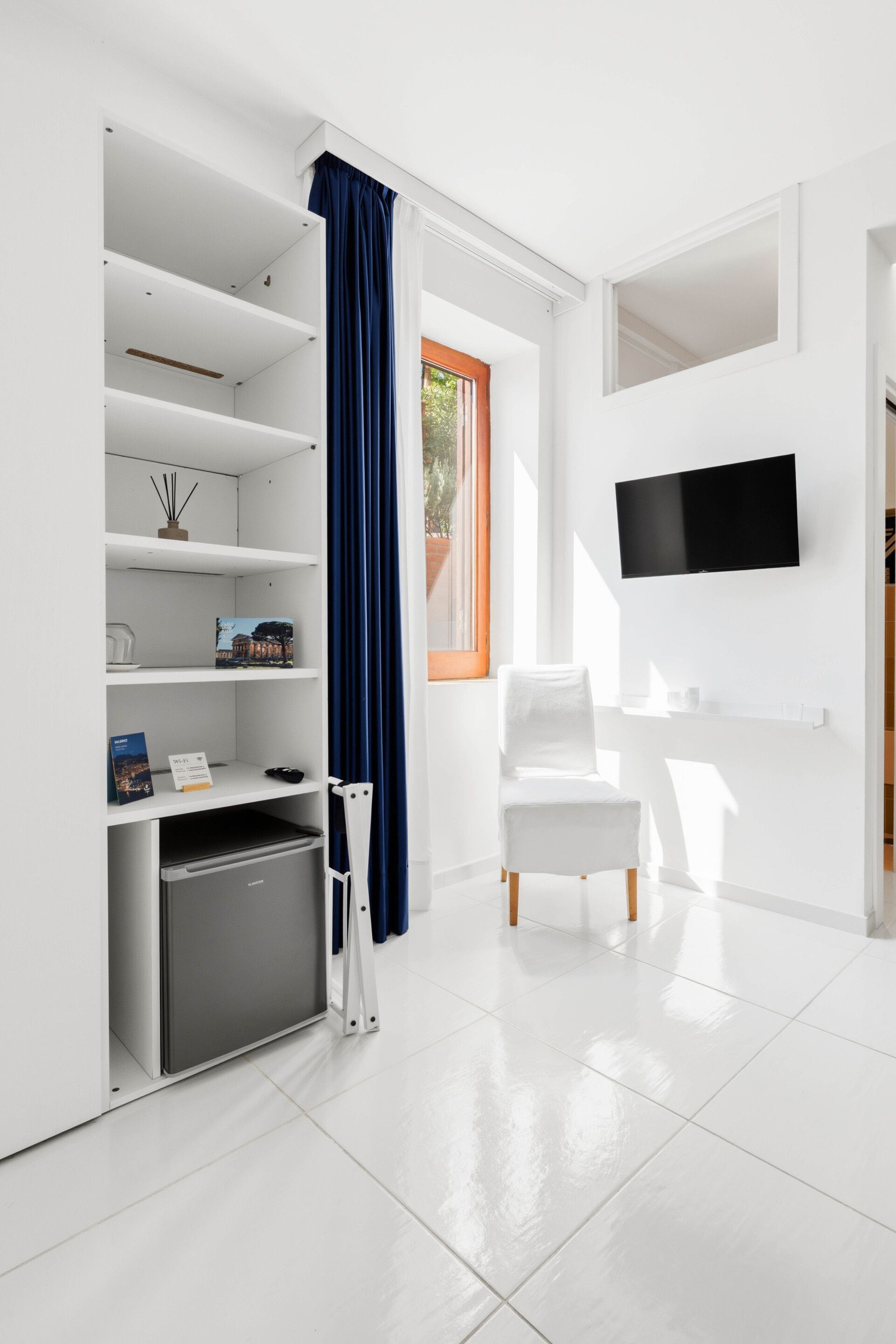 Una camera moderna con arredamento bianco, una TV a schermo piatto montata a parete, una poltrona bianca, un angolo cottura e una finestra con vista sul verde.
