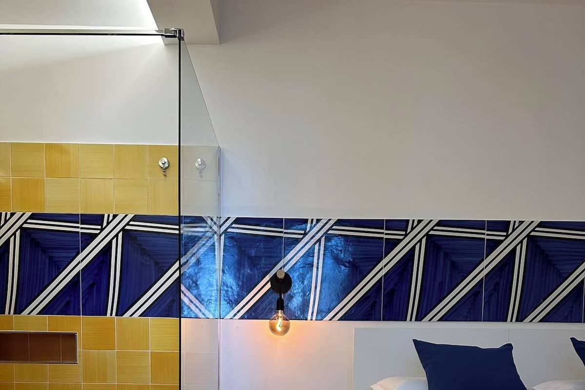 Et moderne soveværelse med et brusekabine i glas, en seng med mørkeblåt sengetøj og et loft med geometrisk mønster.