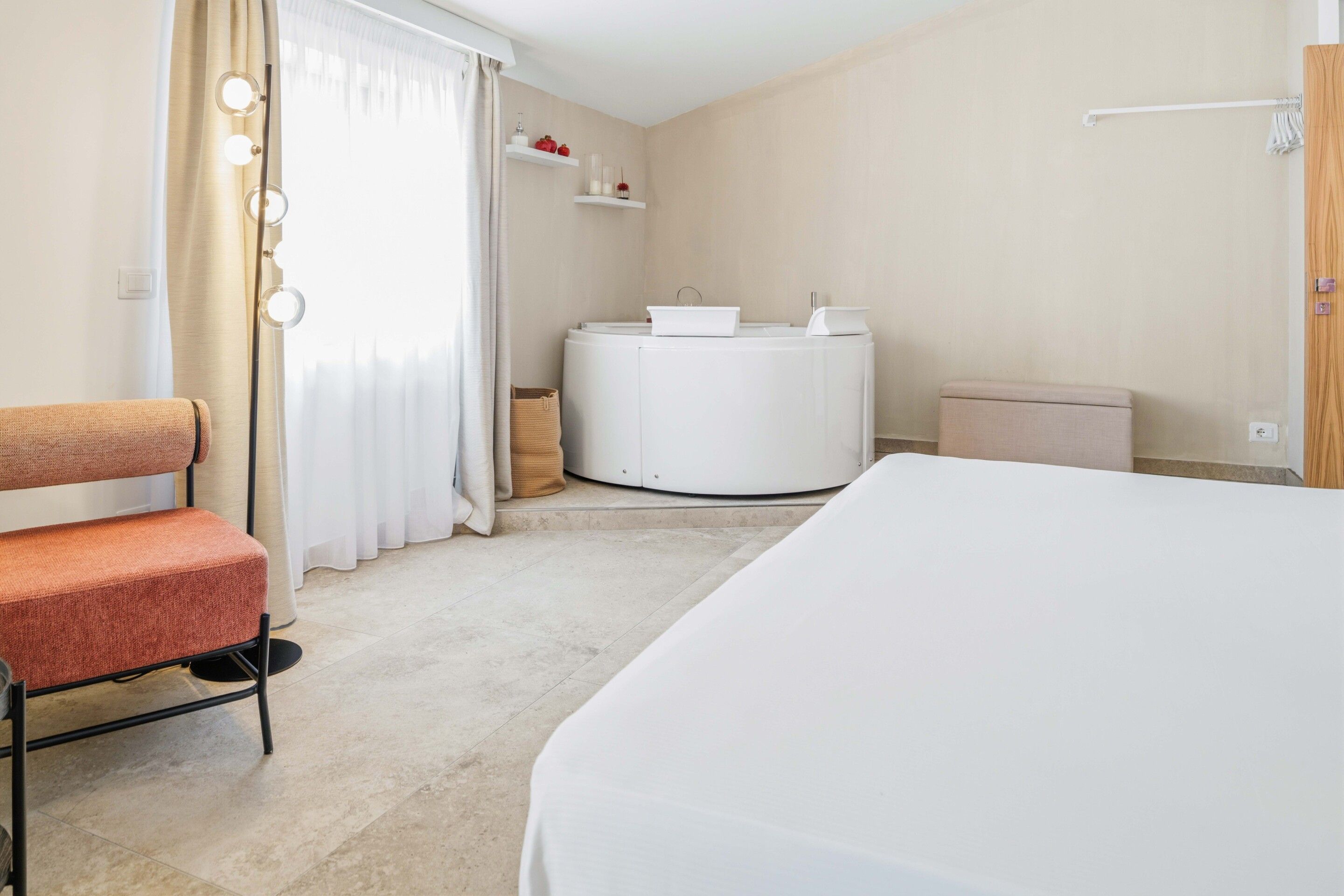 A hotel room with a bed, a chair, a round table with a white tablecloth, a small trash bin, and a wall-mounted shelf with decorative items.