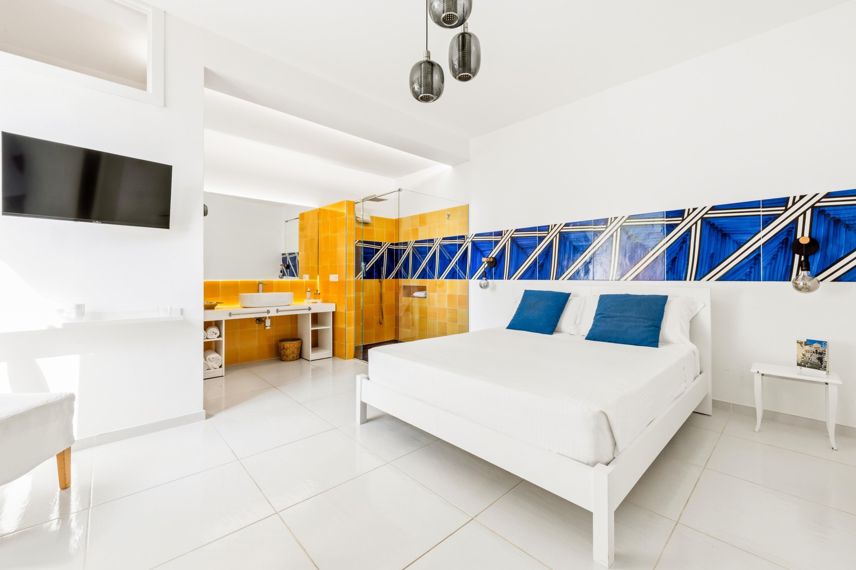 A modern bedroom with a large bed, a TV, a bathroom with yellow tiles, and a blue patterned wall.