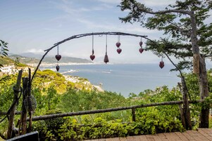 A view of a coastal landscape with a wooden archway adorned with heart-shaped ornaments, leading to a lush green area with cacti and a clear view of the sea and distant hills.