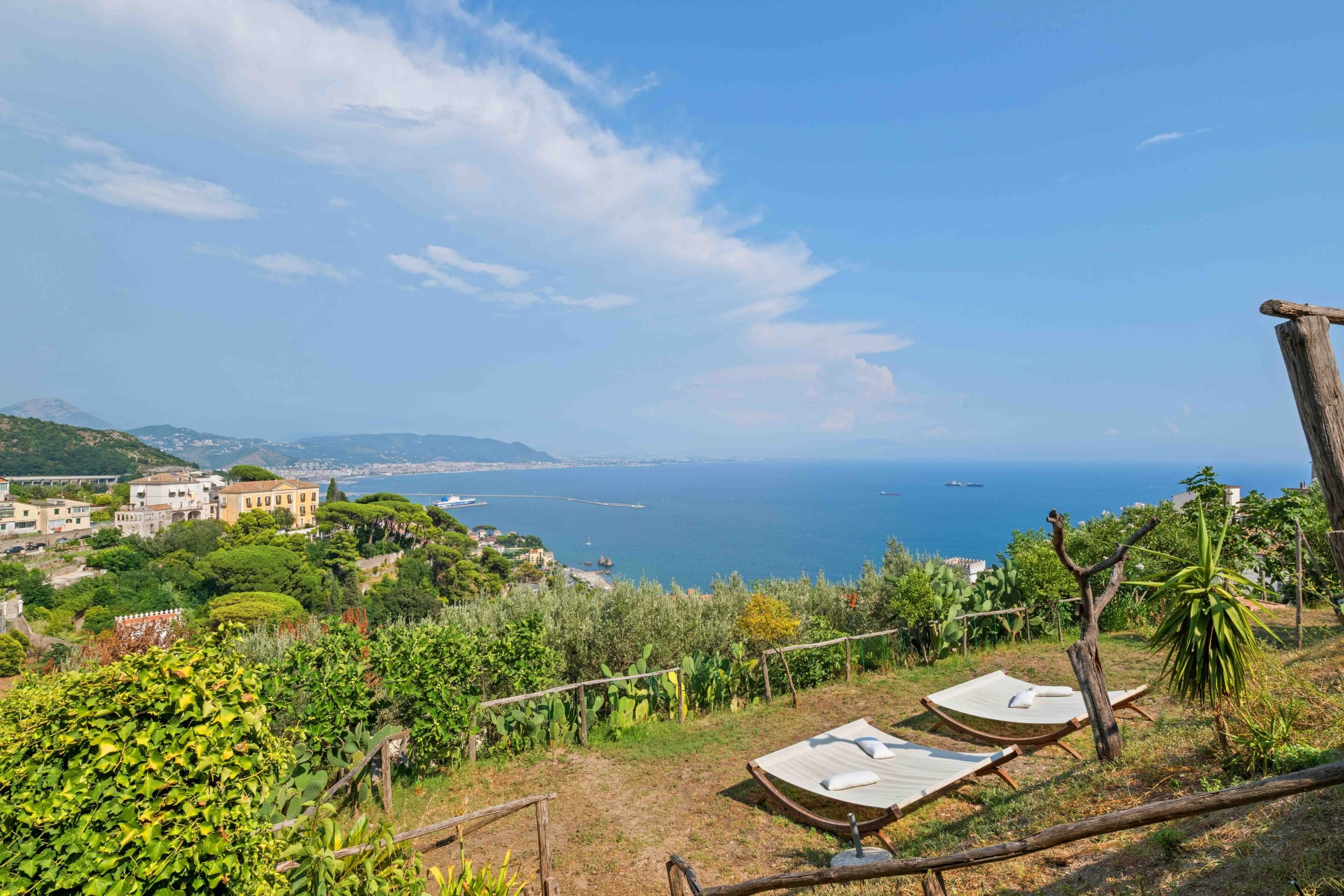 A coastal view with two hammocks overlooking the sea, surrounded by greenery and buildings.