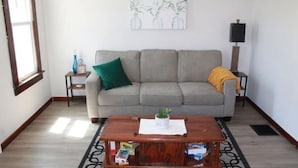 A living room with a grey sofa, a wooden coffee table with a plant, a green pillow, a small side table with a clock, and a wall-mounted TV. - Cozy little cottage in the heart of the Gem City (Quincy)
