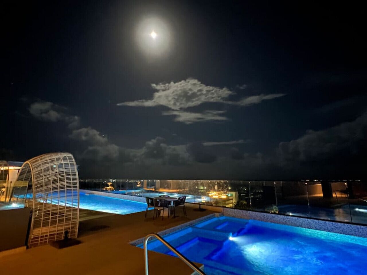 Uma área de piscina no terraço com guarda-corpo de vidro, espaço para sentar e vista para o horizonte urbano à noite.