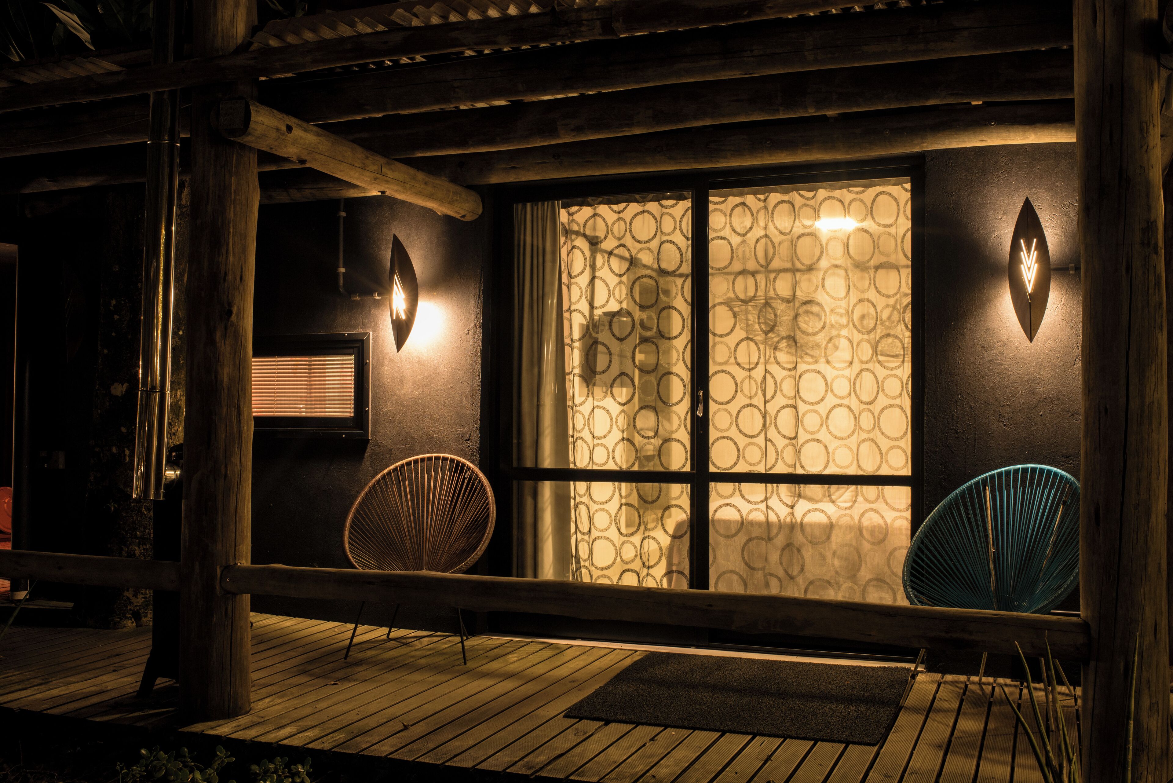 A wooden deck with two chairs, a window with patterned curtains, and wall-mounted lights.