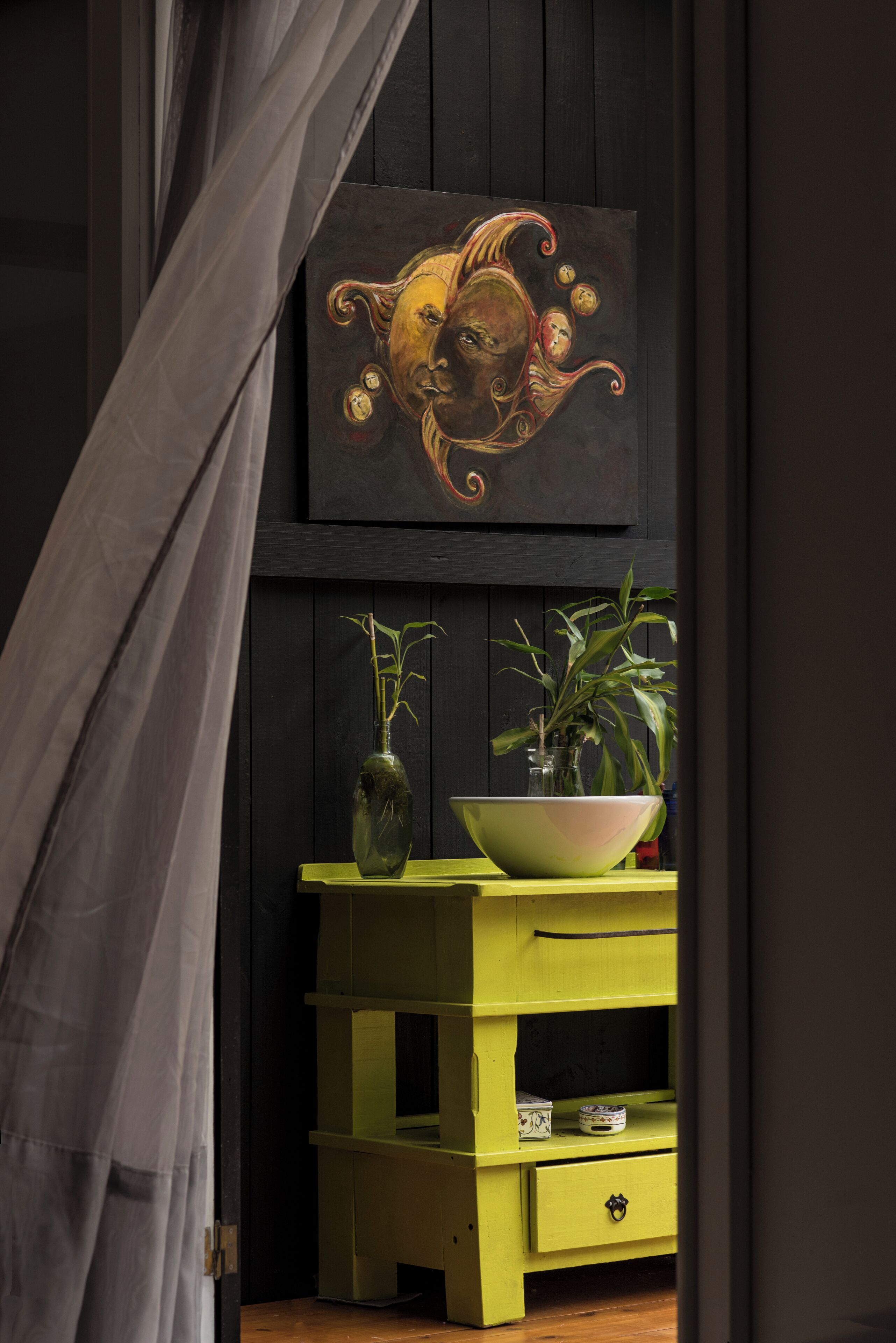 Una habitación con una pared de madera oscura, un aparador verde, un florero con una planta y un cuadro de un rostro.
