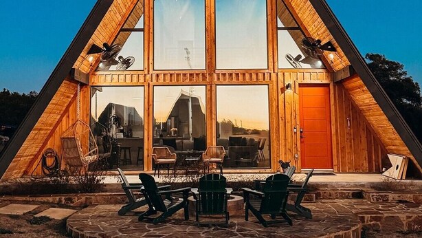 Una cabaña de madera triangular con ventanas amplias, un patio de piedra y mobiliario exterior.