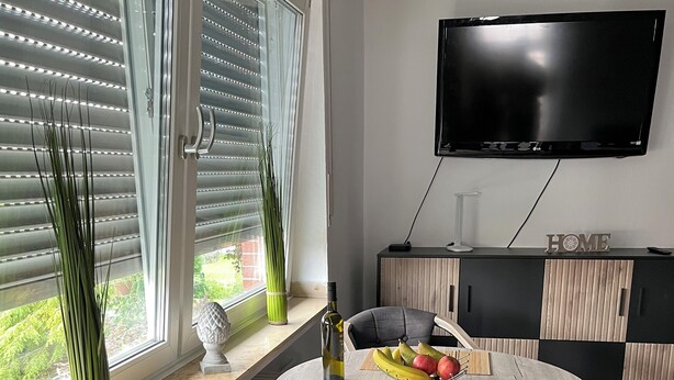 A modern living room with a round dining table, two chairs, a TV mounted on the wall, and a window with blinds. - Apartment 1880 (Werne)