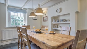 A dining area with a wooden table, chairs, and pendant lights. There is a window with a view of trees, a radiator, and a shelf with kitchen items. - Dollars Cottage - 4 Bed - Llangennith (Swansea)
