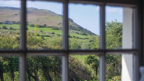 A view through a window of a green landscape with hills and a house in the distance. - Dollars Cottage - 4 Bed - Llangennith (Swansea)