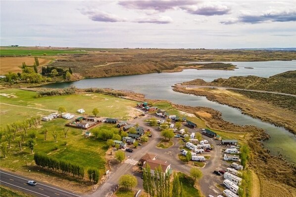 Vista aérea de un camping con vehículos recreativos estacionados a lo largo de una carretera, rodeado de campos abiertos y una masa de agua.