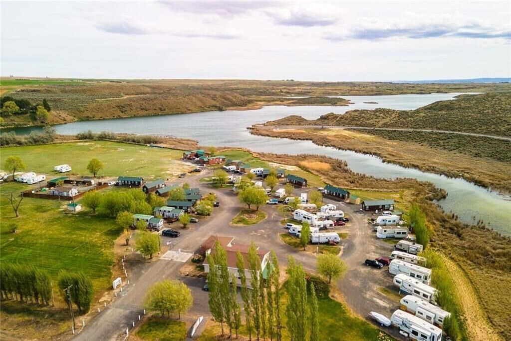 Un terrain de camping pour véhicules récréatifs, avec plusieurs véhicules stationnés dans des zones aménagées, un bâtiment central et une rivière en arrière-plan.