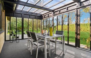 A conservatory with a glass roof, a dining table with chairs, and a view of a garden.