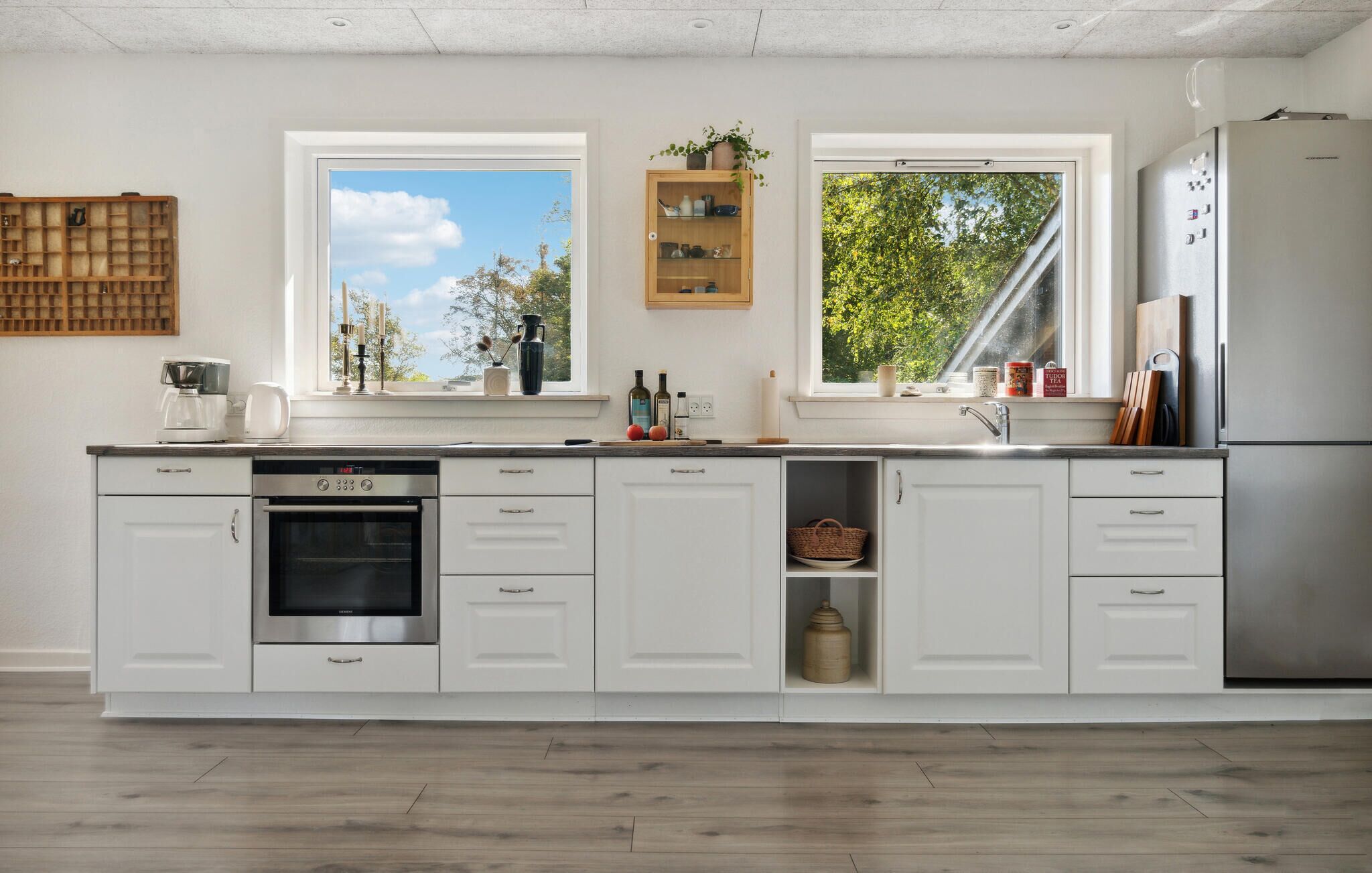 Cocina moderna con armarios blancos, horno empotrado, lavavajillas y refrigerador de acero inoxidable. Dos ventanas ofrecen luz natural y vistas al exterior.