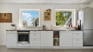A modern kitchen with white cabinetry, a built-in oven, a dishwasher, and a stainless steel refrigerator. There are two windows providing natural light and views of greenery outside. - Awesome home in Nykøbing M (Nykøbing M)