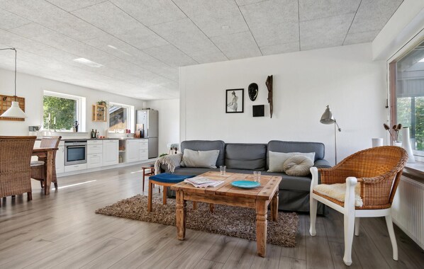 A spacious living area with a grey sofa, a wooden coffee table, and wicker chairs. A kitchen with white cabinets and a window is visible in the background.