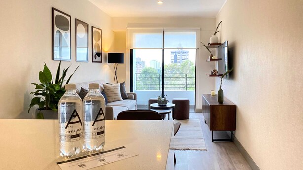 A modern living room with a sofa, a coffee table, and a TV mounted on the wall. There are two bottles of water on the table, and a plant in the corner. - Wynwood The Classic in San Isidro (Lima)