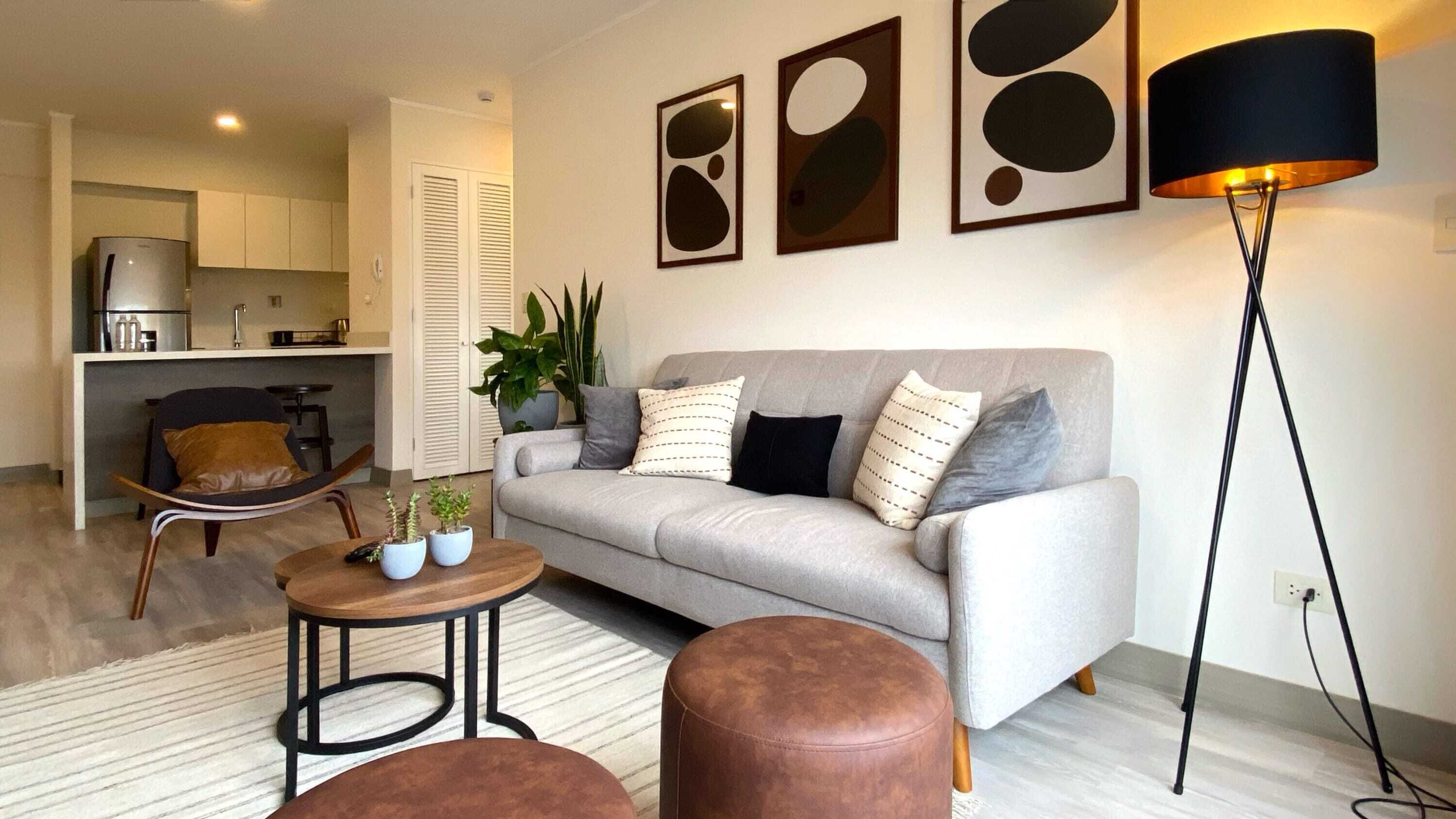 A living room with a grey sofa, two brown ottomans, a small round table with two potted plants, a floor lamp, and abstract art on the wall.