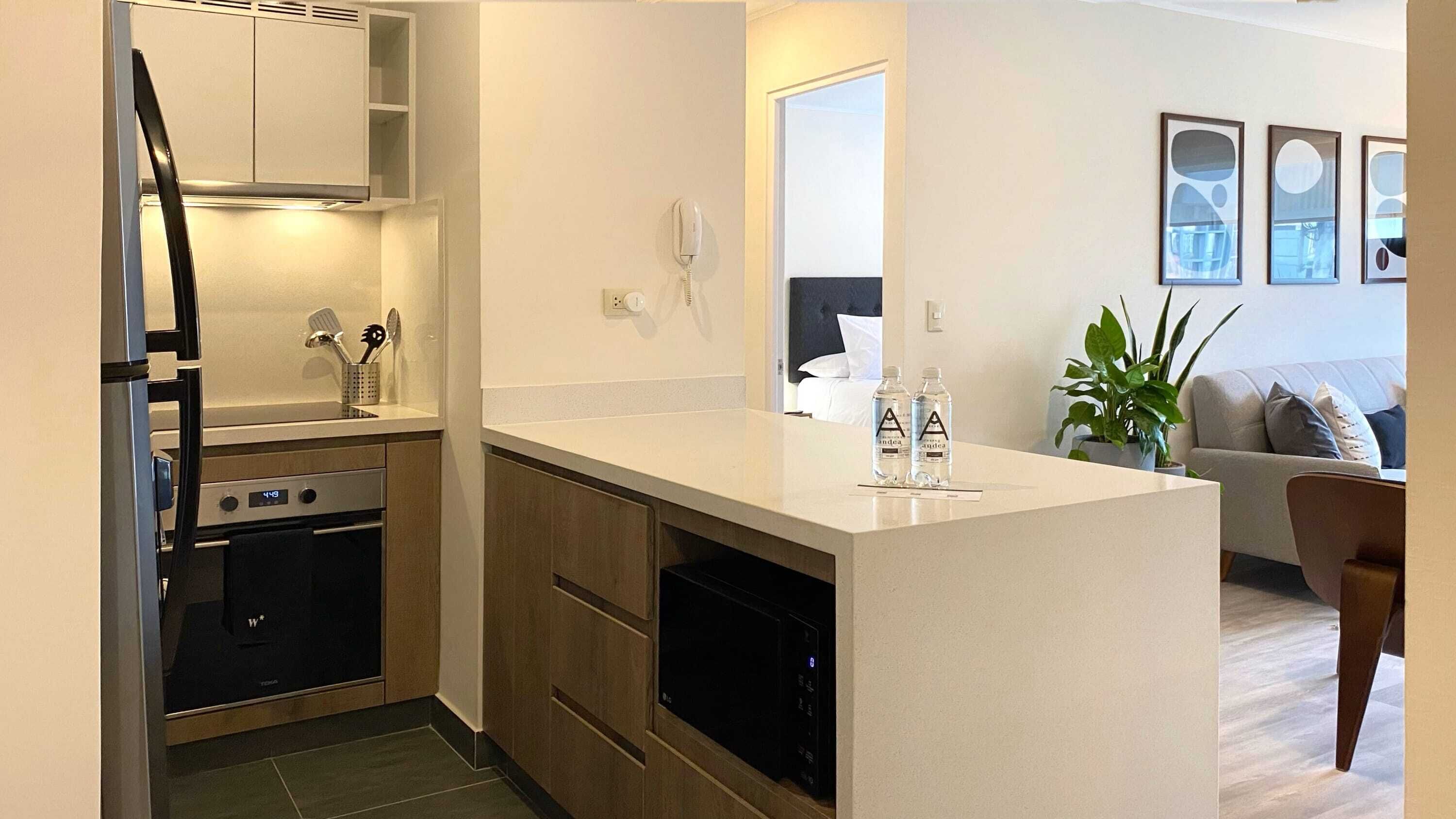 Eine moderne Küche mit integriertem Ofen, Mikrowelle und Geschirrspüler. Eine kleine Kücheninsel beherbergt ein Spülbecken und einige Wasserflaschen. Angrenzend befindet sich ein Wohnbereich mit einer Couch und einer Pflanze.