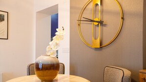 A dining area with a round table, chairs, a vase with flowers, and a wall-mounted clock. - Villa Pupin SARAISTE (Ohrid)