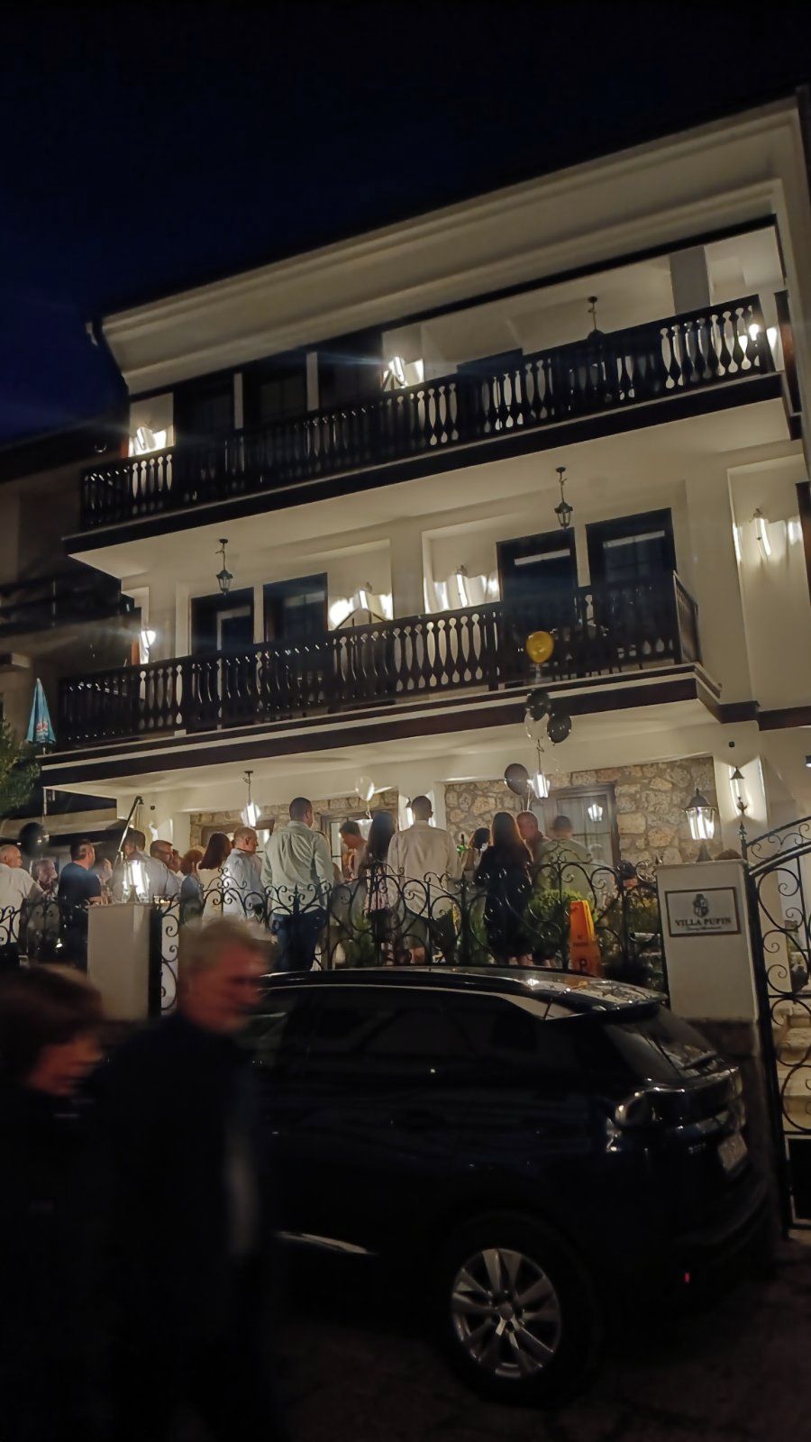 Un edificio de varios pisos con balcones, un coche estacionado frente a él y un grupo de personas reunidas afuera.