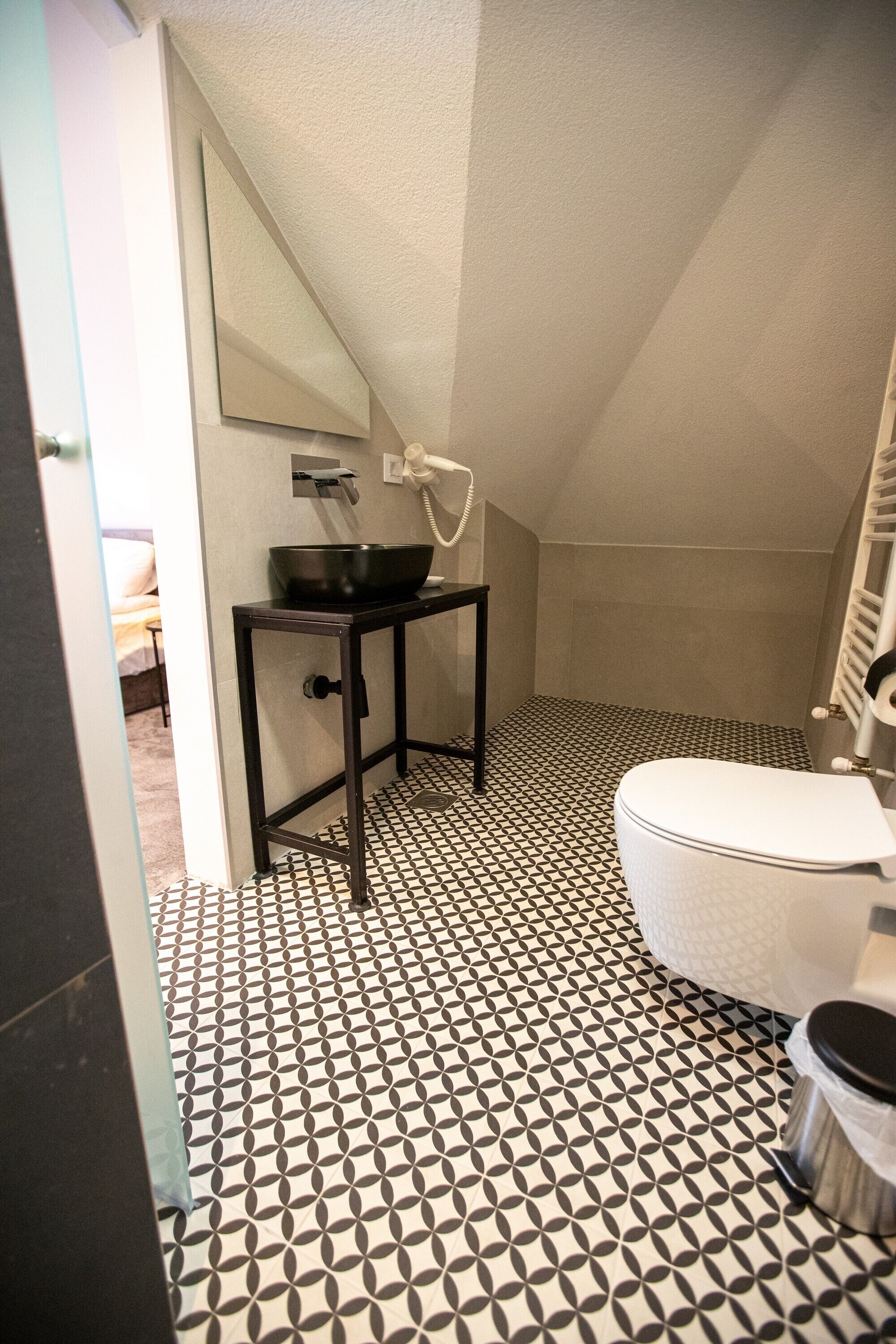 Um banheiro com piso em padrão preto e branco, vaso sanitário branco, pia preta com espelho acima e área de chuveiro.