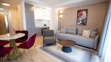 Un salon moderne avec un canapé gris, des fauteuils rouges, une table basse ronde et une cuisine en arrière-plan.