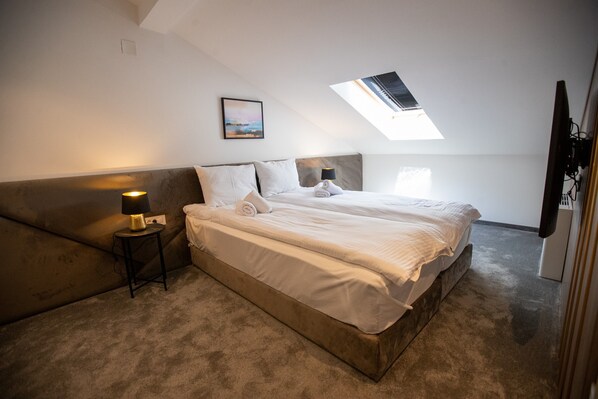 A bedroom with a large bed, a small bedside table with a lamp, a picture on the wall, and a skylight above the bed. - Villa Pupin SARAISTE (Ohrid)