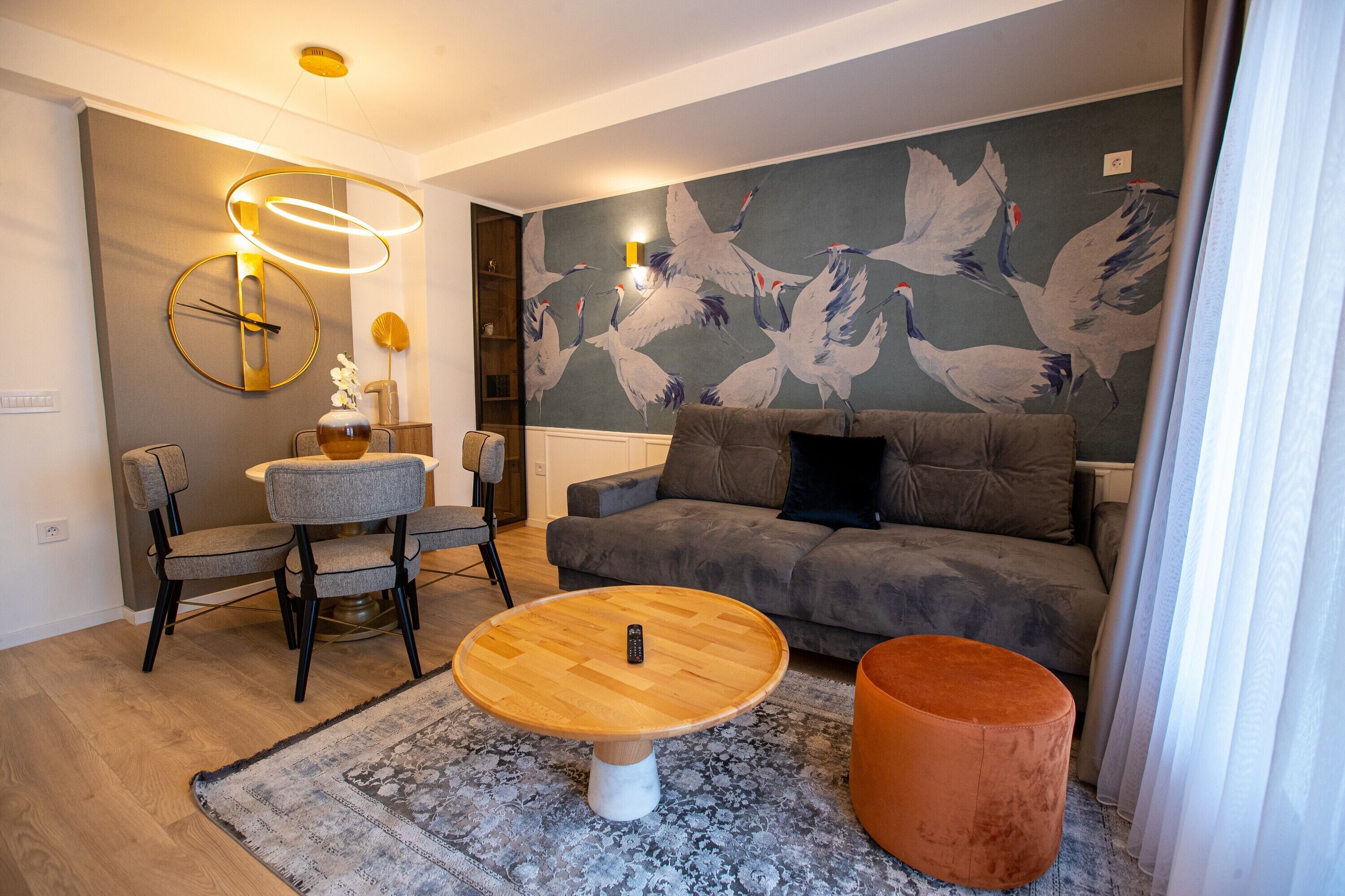 A living room with a grey sofa, a round wooden coffee table, a grey armchair, a small orange ottoman, and a wall with a bird mural.